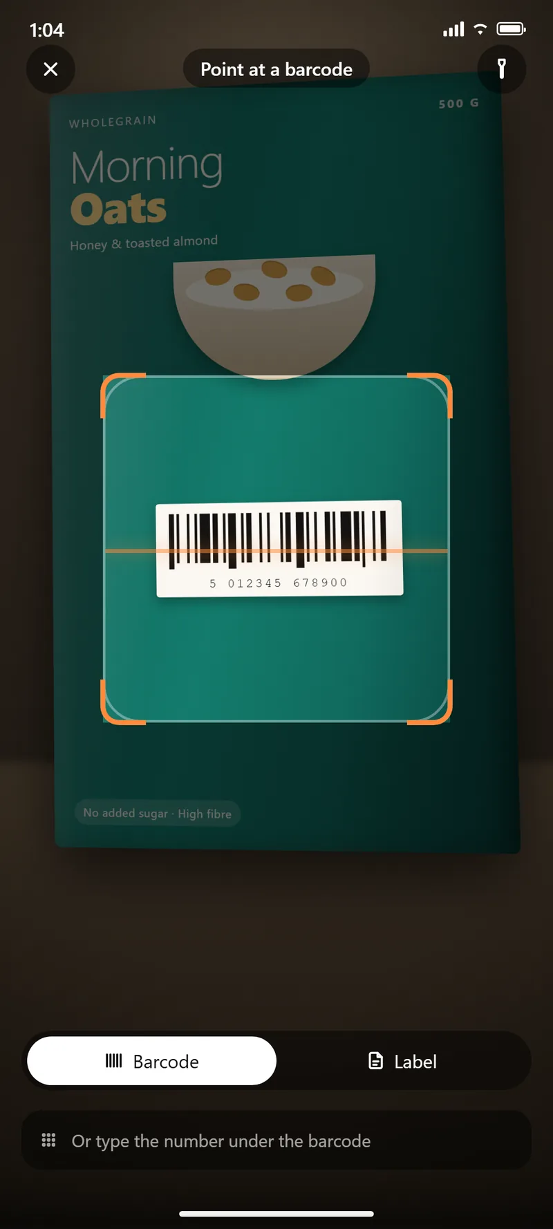 The scanner in barcode mode, aimed at the barcode on a box of Morning Oats cereal standing on a counter. An orange framing bracket surrounds the barcode, with a Barcode and Label toggle and a field to type the number under the barcode below.